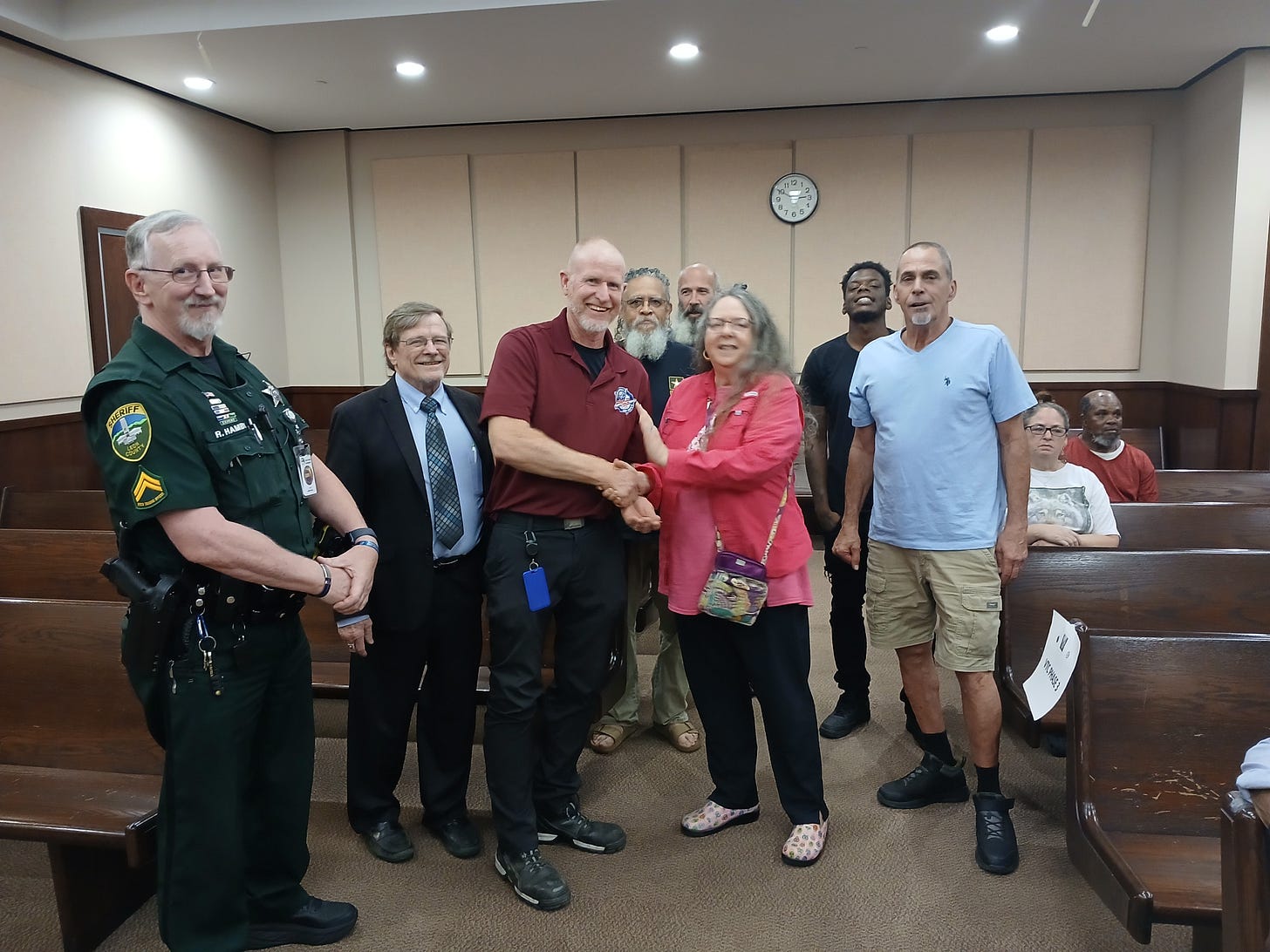 The image shows a group of people standing together and smiling in what appears to be a courtroom or a meeting room with wooden benches. In the front center, a man in a maroon shirt is shaking hands with a woman in a pink top. To the left, a sheriff in uniform stands with them, and a man in a suit and tie is next to the sheriff. Several other men and women are in the background, some standing and some sitting on the benches. A clock on the wall shows the time as 9:24. The atmosphere is positive and friendly, with several people smiling for the photo. No recognizable figures, landmarks, or brands are clearly identifiable in this image. The image shows a group of people standing together and smiling in what appears to be a courtroom or a meeting room with wooden benches. In the front center, a man in a maroon shirt is shaking hands with a woman in a pink top. To the left, a sheriff in uniform stands with them, and a man in a suit and tie is next to the sheriff. Several other men and women are in the background, some standing and some sitting on the benches. A clock on the wall shows the time as 9:24. The atmosphere is positive and friendly, with several people smiling for the photo. No recognizable figures, landmarks, or brands are clearly identifiable in this image.