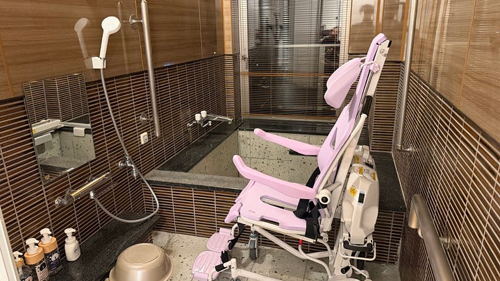 Image 1 alt text: An accessible bathroom with a natural stone soaking tub, grab bars, handheld shower, and a pink mechanized shower chair lift positioned beside the bath for wheelchair transfers.  Image 2 alt text: A spacious Japanese-Western style guest room with twin beds, warm lighting, wood furniture, and a raised tatami platform decorated with a floral arrangement.