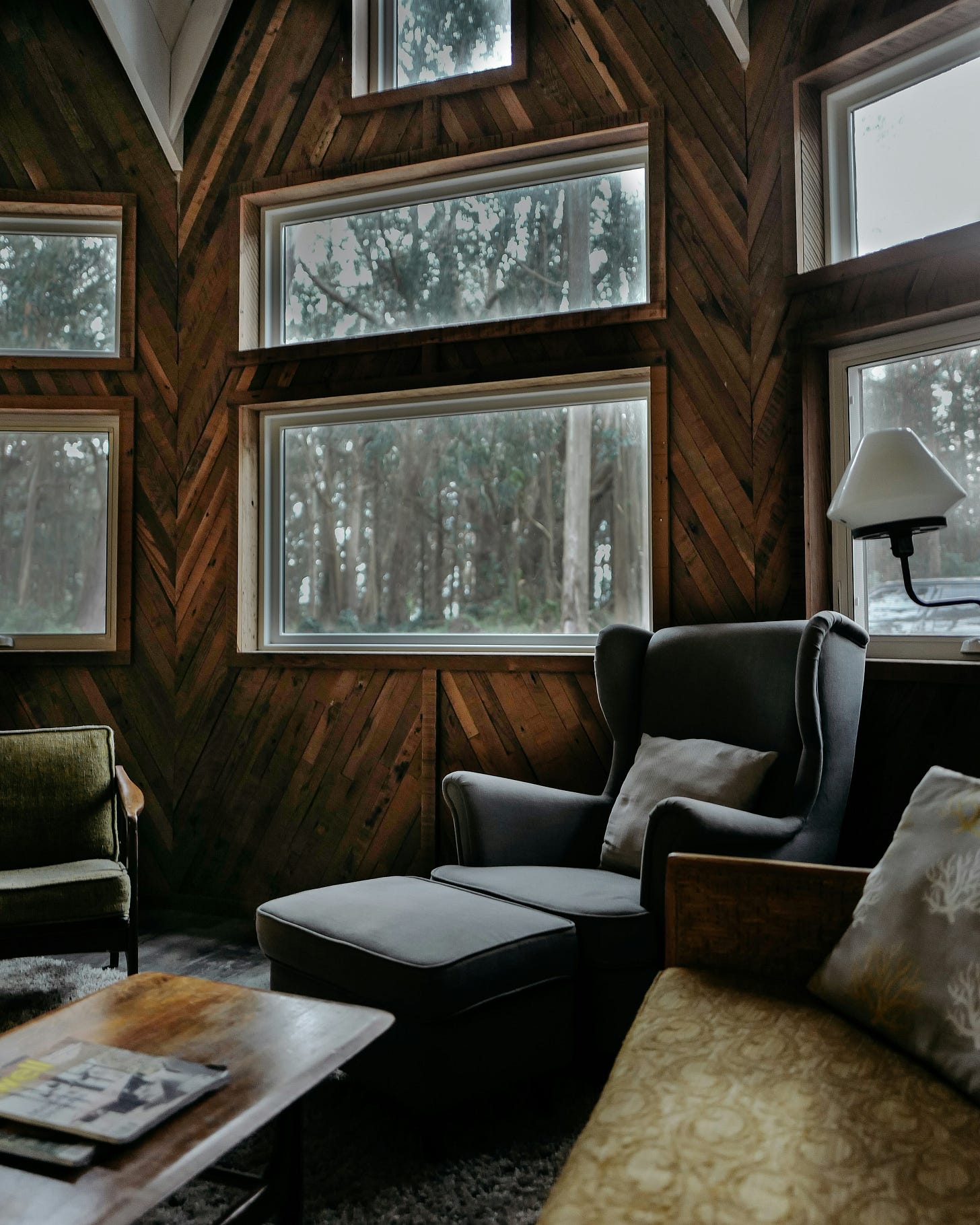 A grey chair and footrest with an old pillow sit inside a cozy wood cabin room. The forest is visible outside A grey chair and footrest with an old pillow sit inside a cozy wood cabin room. The forest is visible outside