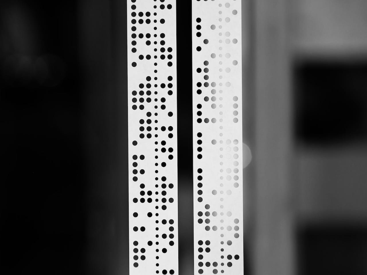 Two vertical strips of punched tape with rows of circular holes used for data encoding for an early computer, set against a blurred dark and light background.
