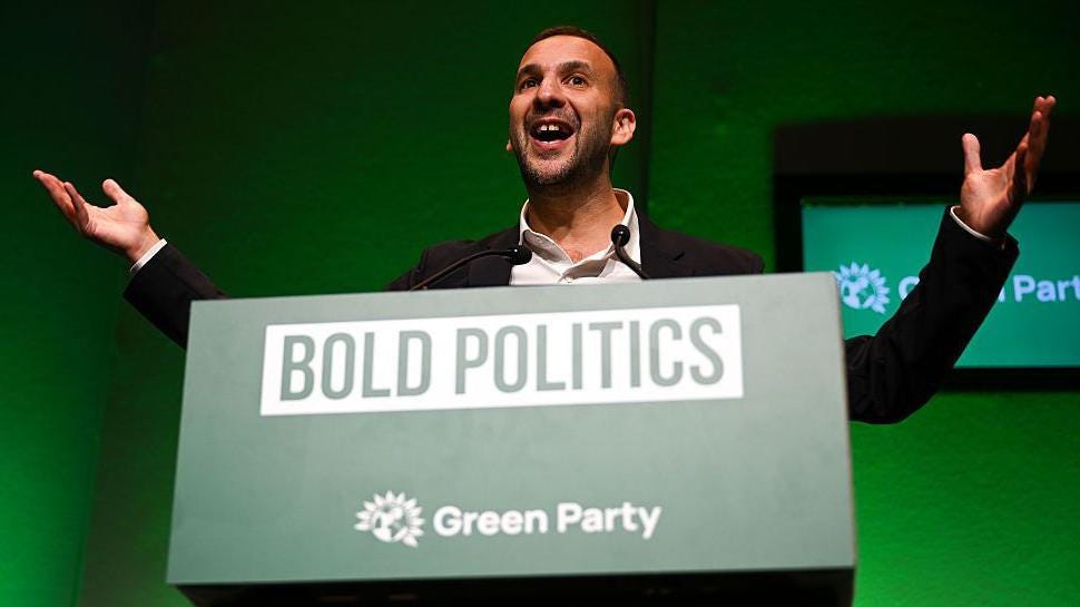 Green Party leader Zack Polanski stands with his arms outstretched as he delivers his speech on the first day of the Green Party Conference in front of a sign saying "bold politics".