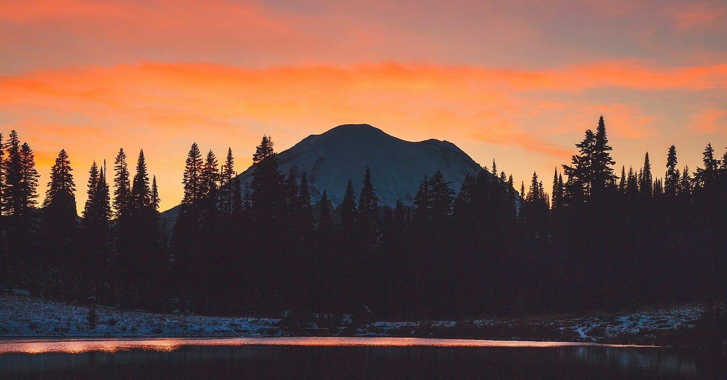 A snow-covered mountain and dark tree line silhouetted against a vivid orange and pink sunset sky, with a calm lake reflecting the colors below.