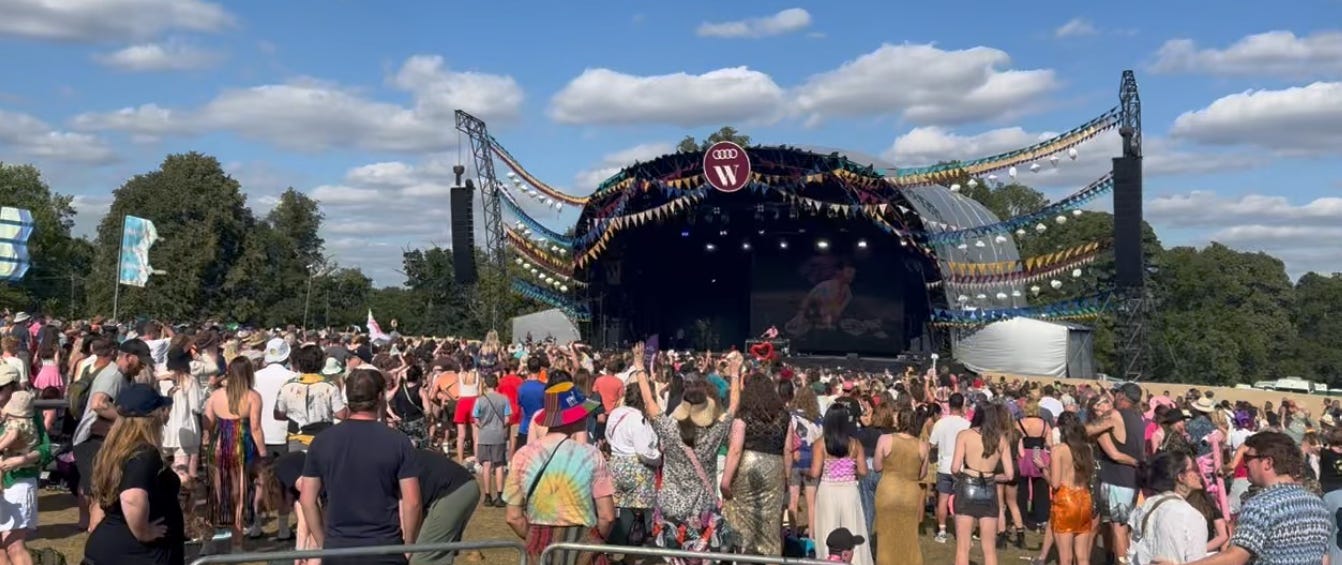 A large outdoor festival crowd gathered in front of a brightly decorated stage with colourful banners and a circular W sign. Trees surround the area and the sky is partly cloudy.
