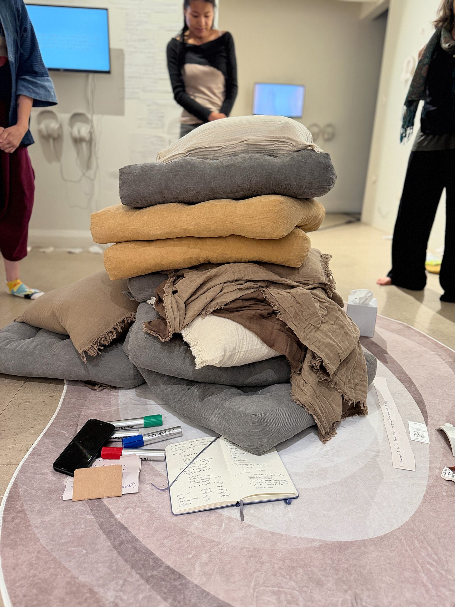 A stack of folded throws, blankets, and cushions piled on the round rug. A few objects rest on the floor beside them. In the background, a monitor on the wall and people standing.