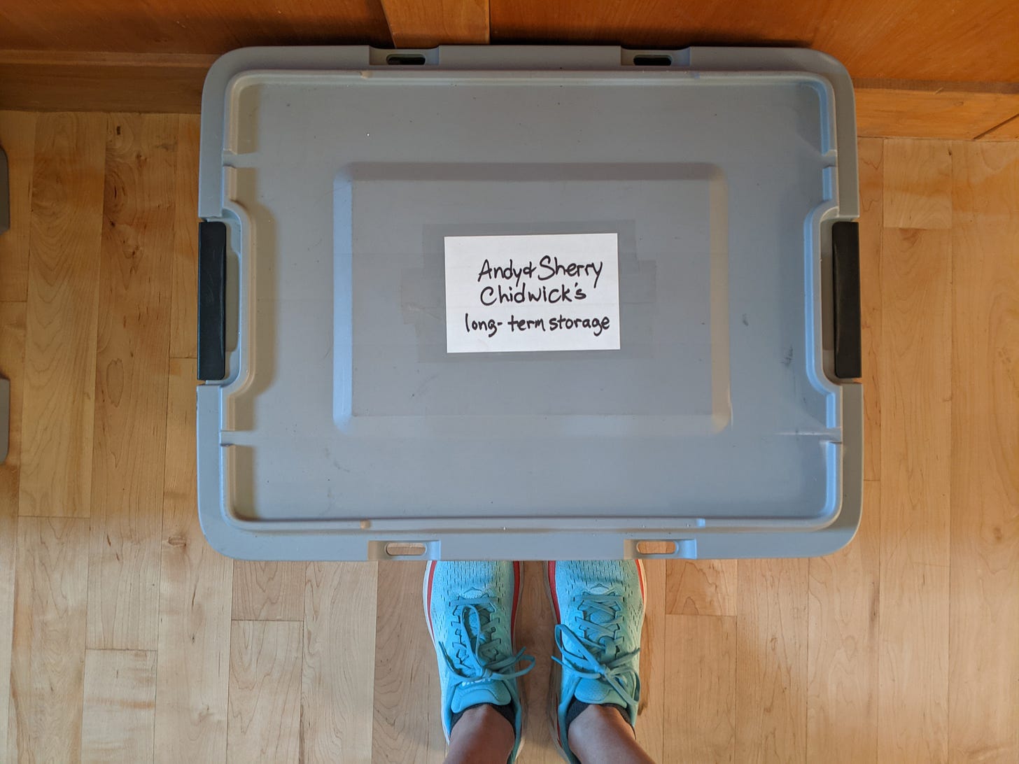 plastic tub labeled Andy & Sherry Chidwick's long-term storage, sitting on a hardwood floor in front of a pair of feet in blue running shoes plastic tub labeled Andy & Sherry Chidwick's long-term storage, sitting on a hardwood floor in front of a pair of feet in blue running shoes