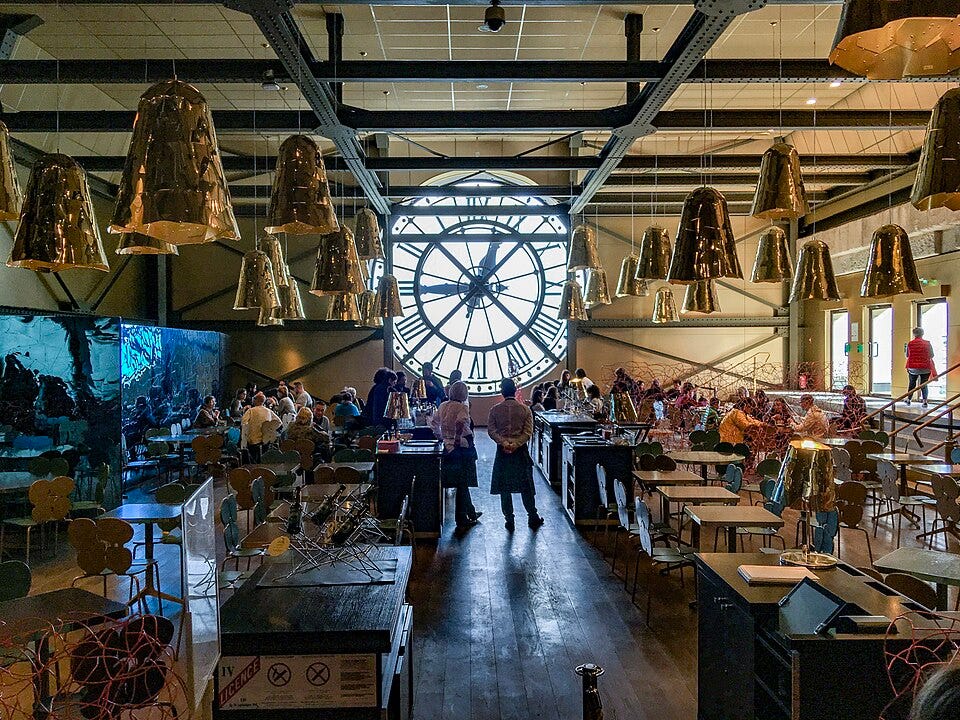 Diners sit at café tables beneath a huge backlit clock at the Musée d’Orsay, while two apron-wearing staff members stand in the central aisle. Diners sit at café tables beneath a huge backlit clock at the Musée d’Orsay, while two apron-wearing staff members stand in the central aisle.
