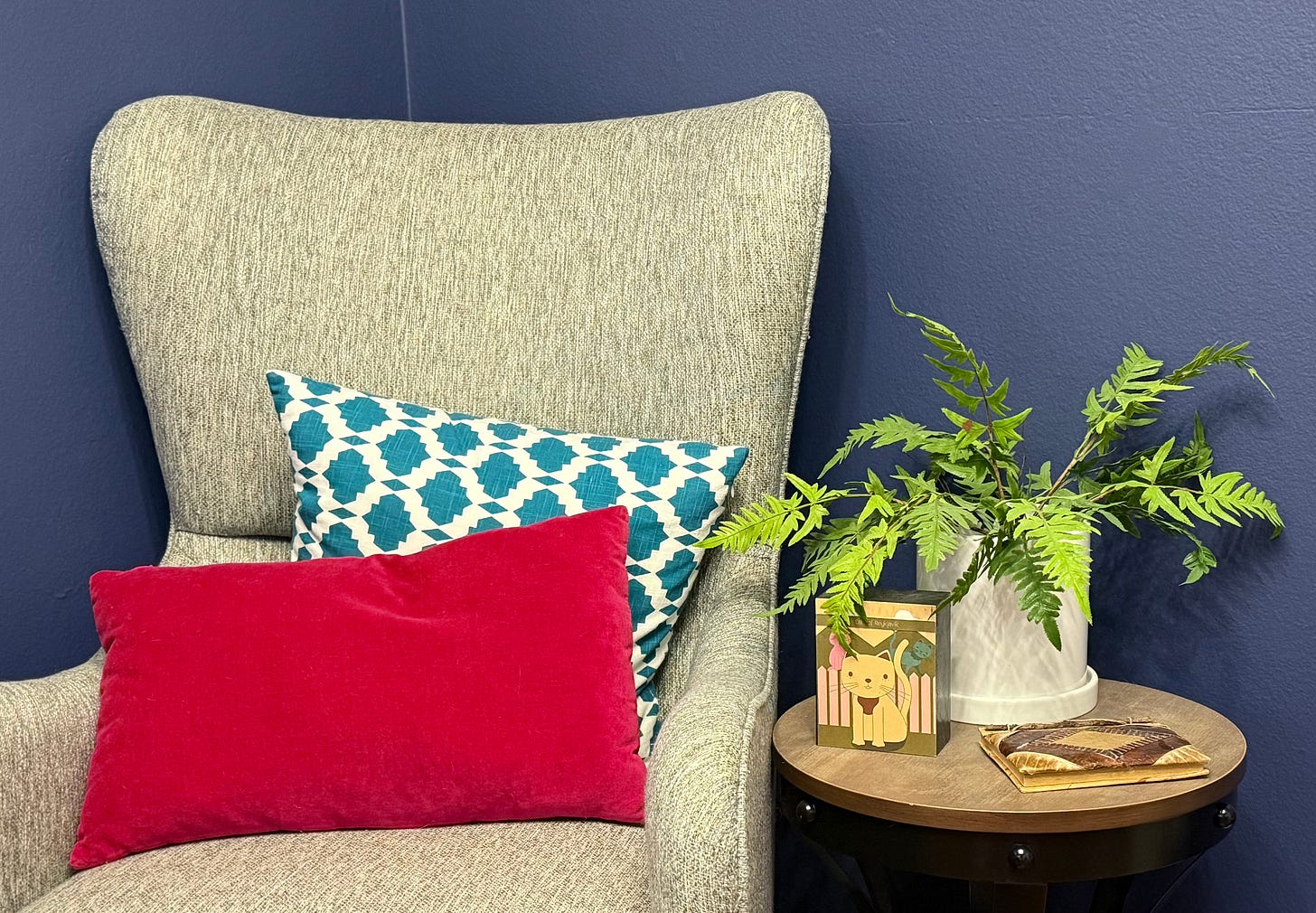 An empty gray armchair with red and teal pillows, next to an end table with a potted fern, a journal, and a drawing of a cat