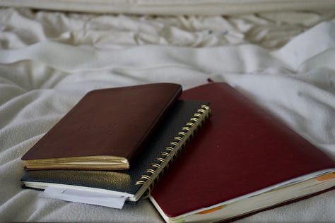 A collection of moody, low-lit photographs including a carefully arranged writing table with tea and ink, stacked journals on rumpled sheets, and a contemplative silhouette at sunset. The images balance structure and softness, suggesting ritual, reflection, and quiet becoming.