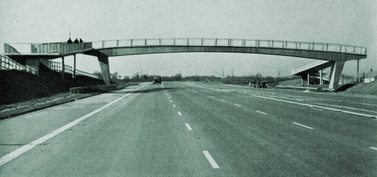 Black and white photograph of a slightly arched concrete footbridge over a motorway that is nearly complete. Some sections of white line are still missing and some construction materials are strewn on the road