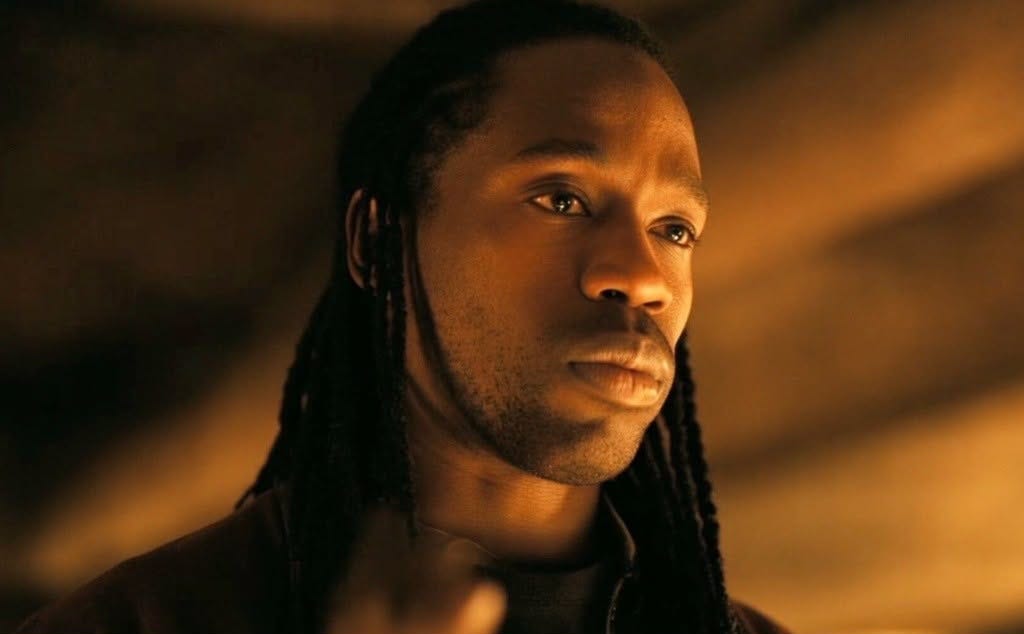 Movie still from The Odyssey. Travis Scott looks on solemnly as the Bard