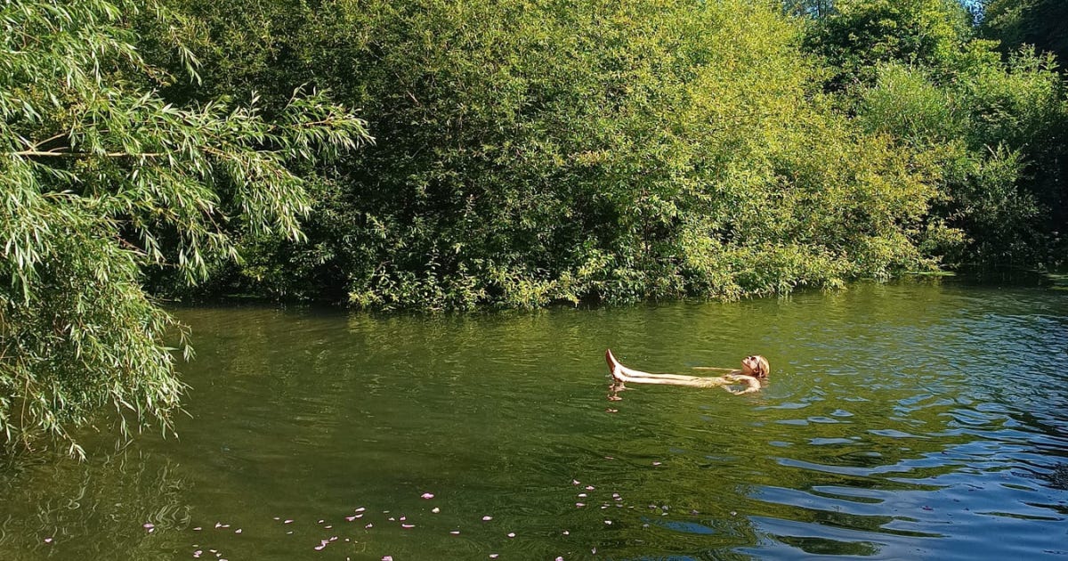A woman lying on her back in the middle of a river surrounded by trees. The sun is shining.