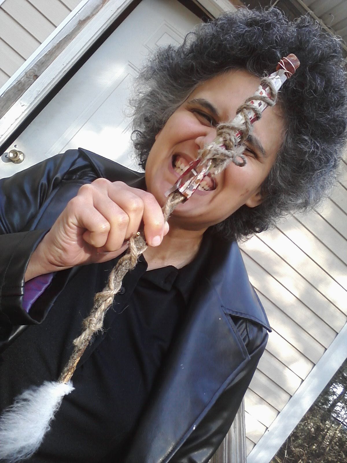 Vas with curly dark gray hair, grinning, holding a simple wrapped wand and wearing a black pleather jacket in front of a door.