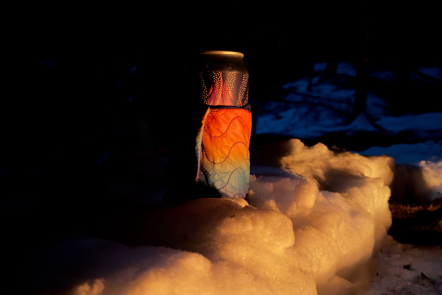 A beer from Blaze Brewing, in a Bevy Sack from Rumpl, sits in a snowbank on a dark night, illuminated by the orange glow of a campfire. A beer from Blaze Brewing, in a Bevy Sack from Rumpl, sits in a snowbank on a dark night, illuminated by the orange glow of a campfire.