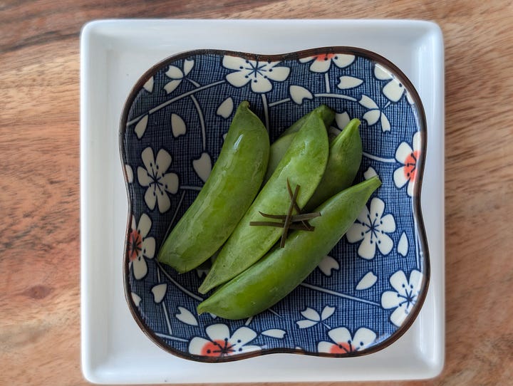 Sheets of kombu seaweed on a wooden cutting board, showing its dark, textured surface, alongside a dish of bright green pickled snap peas with kombu, served in a decorative bowl.