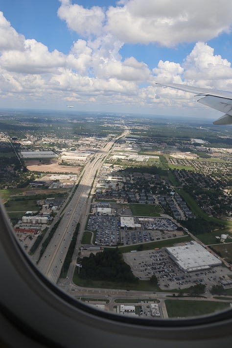Top left - from the airplane window through wisps of cloud to rolling hills. Top middle, from the airplane window, big highway lanes disappear into the distance as the plane banks to land. Top right - bicycle leaning against a pole at a roadside stall. Middle left, small grey house with hedge. Middle middle, dirt road town with two storey shops, wires overhead. Middle right. City square with man leaning looking at the camera with big old building in the background. Cobblestone streets. Bottom left. Close up of bicycle at roadside stop. Road visible with a big blue lorry speeding past. Bottom middle. Most of an orange building with a curved roof and supporting tower wall, graffiti and run down with weeds in the foreground. Bottom right. Big storm clouds over twin railings and city in the background, weeds in the foreground