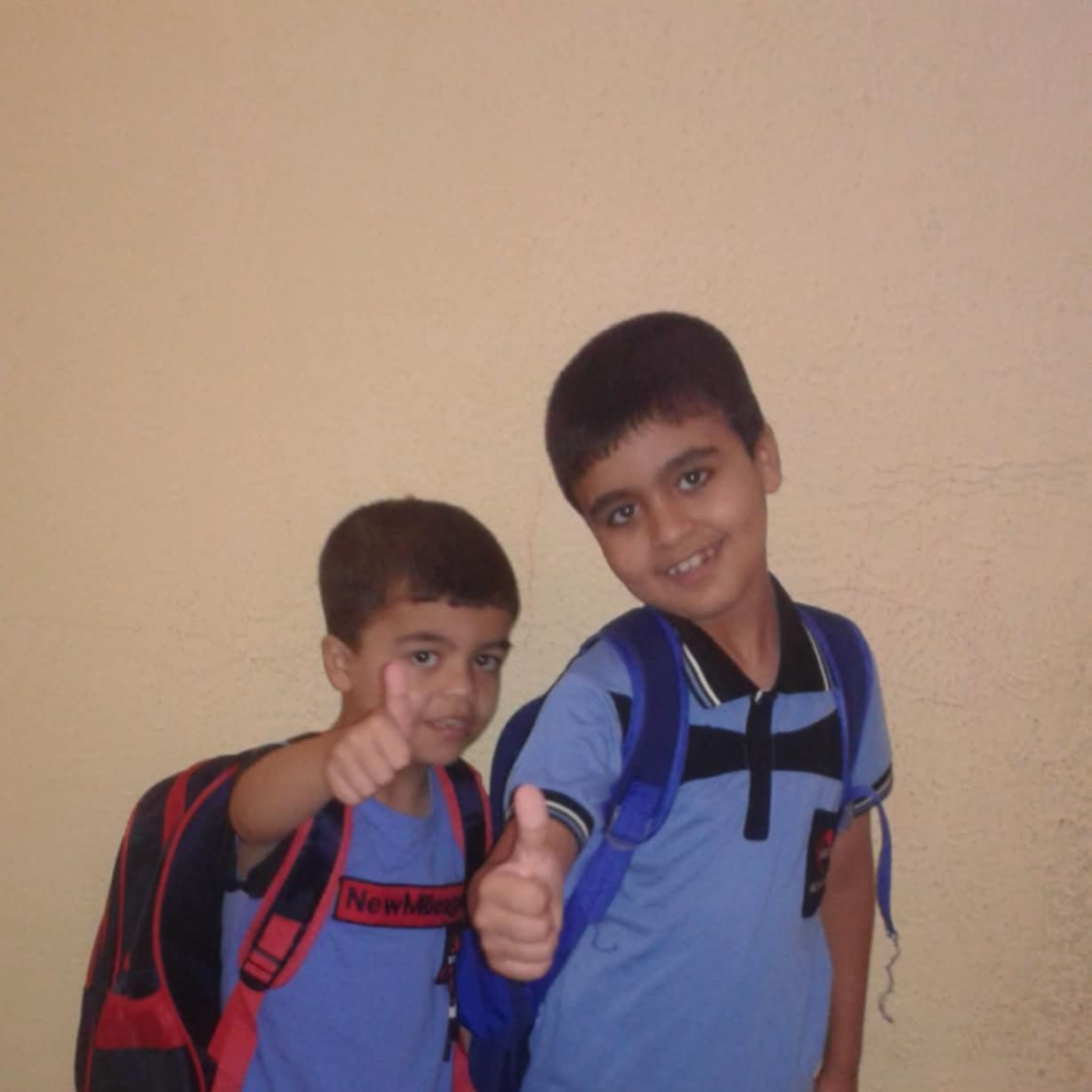 Two young children stand side by side against a plain light-colored wall, both wearing backpacks and blue shirts. They are smiling at the camera and giving thumbs-up gestures, suggesting excitement or confidence, as if ready for school. Two young children stand side by side against a plain light-colored wall, both wearing backpacks and blue shirts. They are smiling at the camera and giving thumbs-up gestures, suggesting excitement or confidence, as if ready for school.