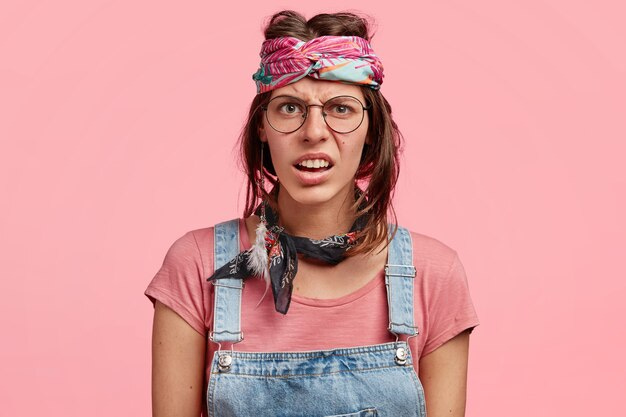 Young white woman wearing huge round glasses, a pink patterned scarf round her forehead that makes her brown hair stick up, denim overalls, a mismatching scarf round her neck, uneven short hair, and an expression that scrunches up her face and puts lines in her forehead. She is intended to look ugly. Young white woman wearing huge round glasses, a pink patterned scarf round her forehead that makes her brown hair stick up, denim overalls, a mismatching scarf round her neck, uneven short hair, and an expression that scrunches up her face and puts lines in her forehead. She is intended to look ugly.