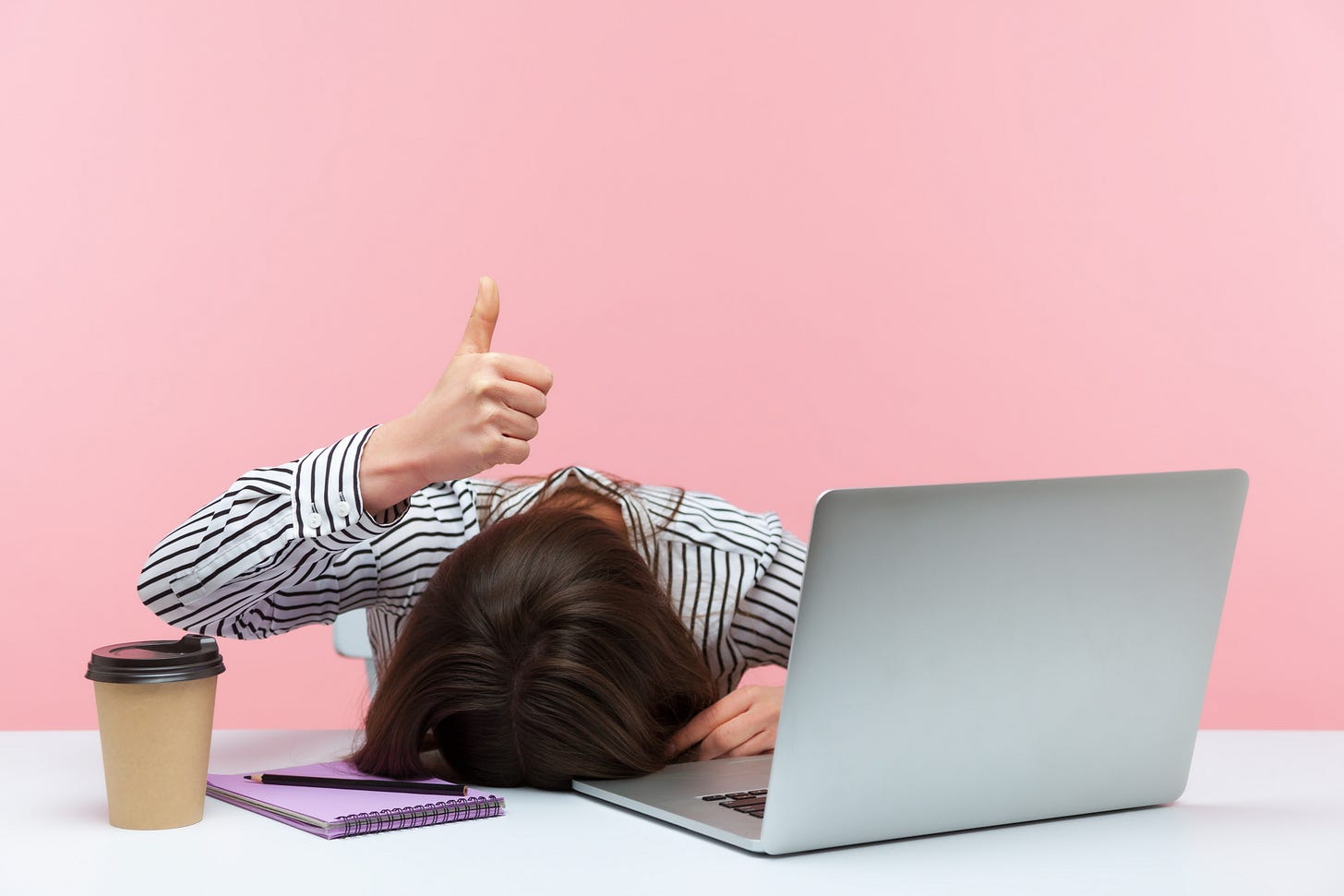 Bored professional woman makes a thumbs up sign while resting her head on her desk