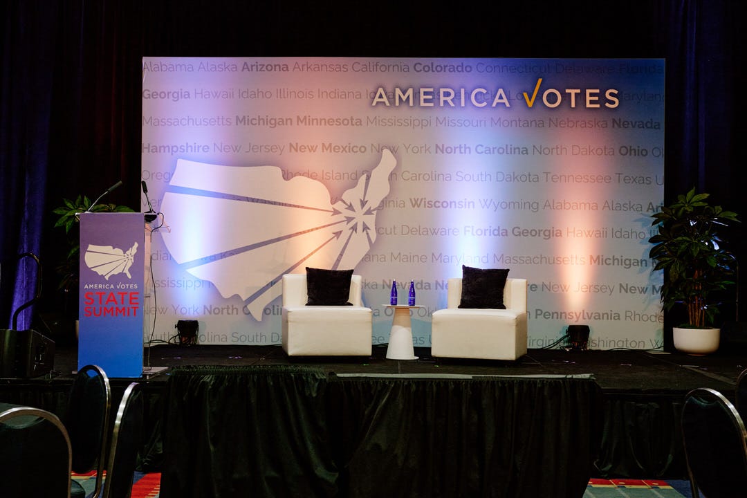 The empty America Votes mainstage with two white chairs and dark pillows, the full state name backdrop lit in blue and gold, before anyone is seated