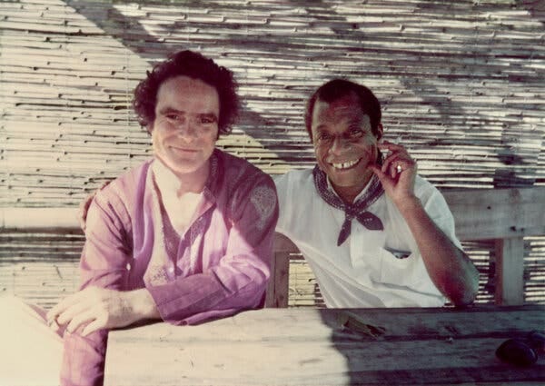 This photograph shows James Baldwin wearing a white polo shirt with a blue bandanna knotted around his neck, a cigarette in his left hand and his right arm draped across the shoulders of a white man with unruly hair and an open-necked lavender shirt.