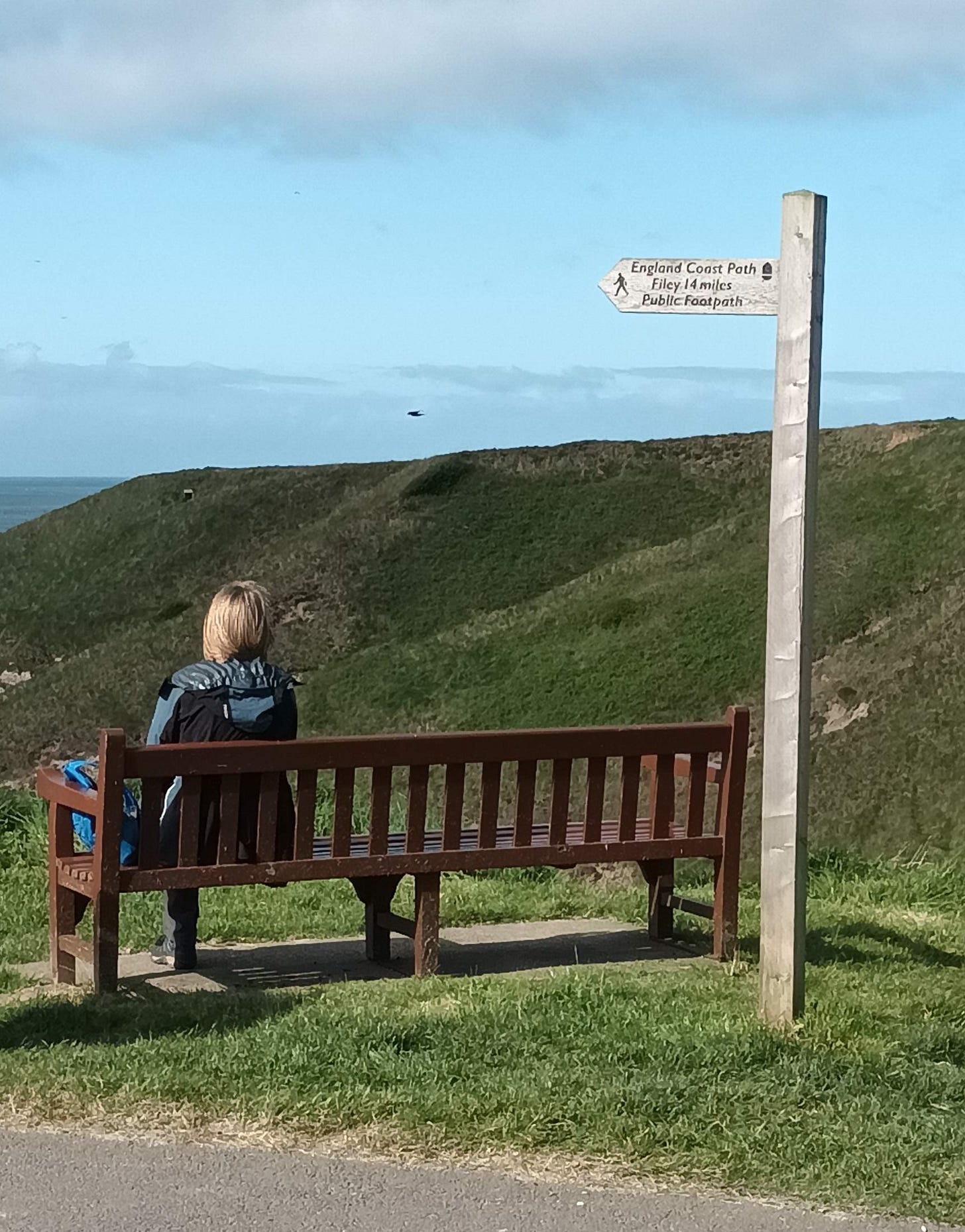 Laura - a blonde woman, sitting on a bench under a sign that says "England Coast Path, Filey, 14 miles, Public Footpath"