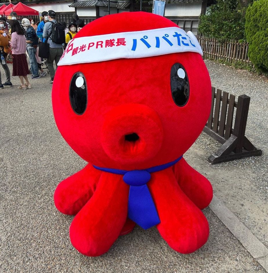 A red octopus mascot with a blue tie and a white headband stands in a park.