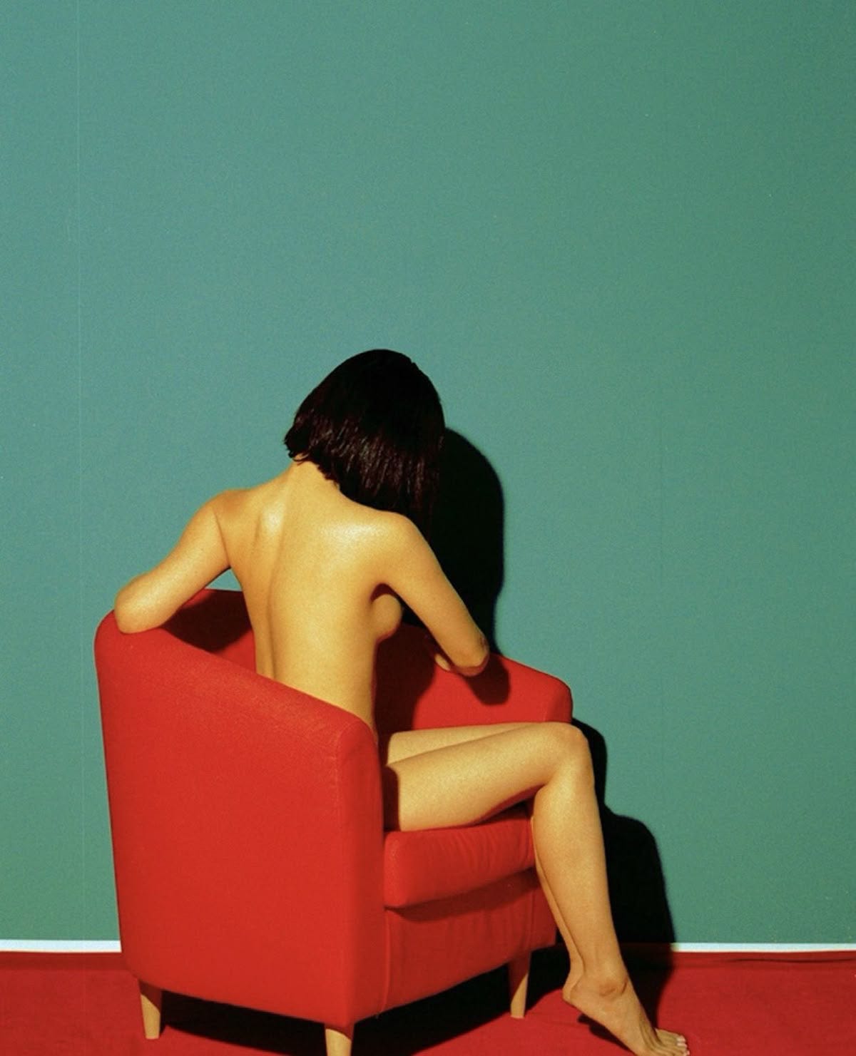 Artistic nude female model seated in a modern red armchair against a teal wall, minimalist contemporary studio photography with bold color contrast.