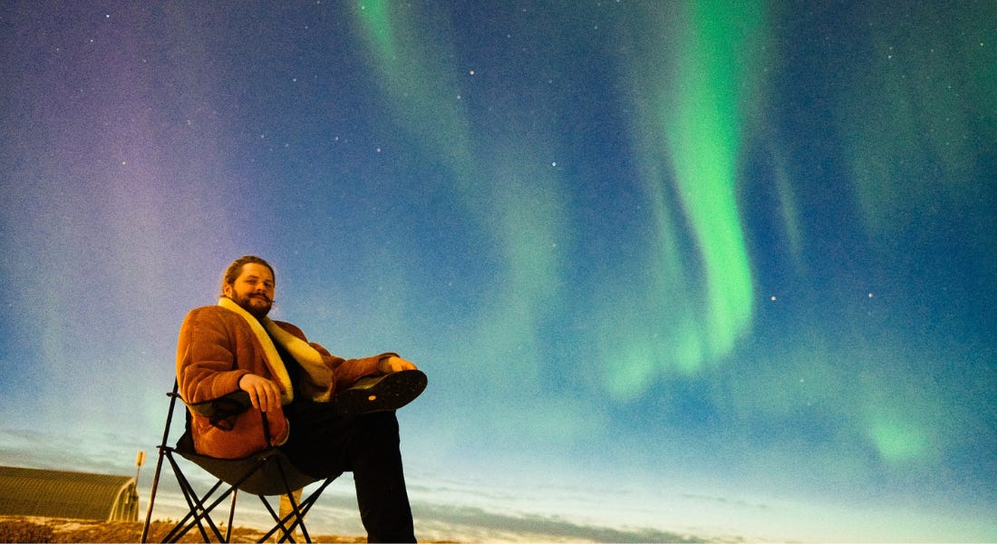 Dan Davison sat on a black chair in front of a night sky filled with Northern Lights