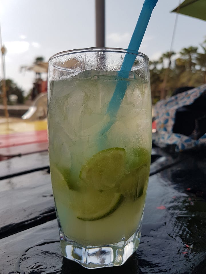 A mojito with mint and lime on a beachside table, next to a small plastic beach bar with outdoor seating set directly on the sand.