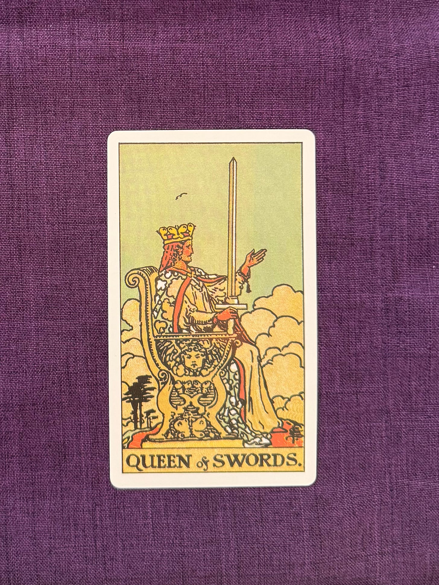 A seated figure sits on an ornate stone throne beneath a sky filled with clouds. The figure wears a crown and flowing robes and holds an upright sword in one hand. The other hand is extended outward with the palm facing up. The throne is decorated with carved motifs, including a winged figure and cloud patterns. Trees and clouds appear in the background, and the figure faces forward.