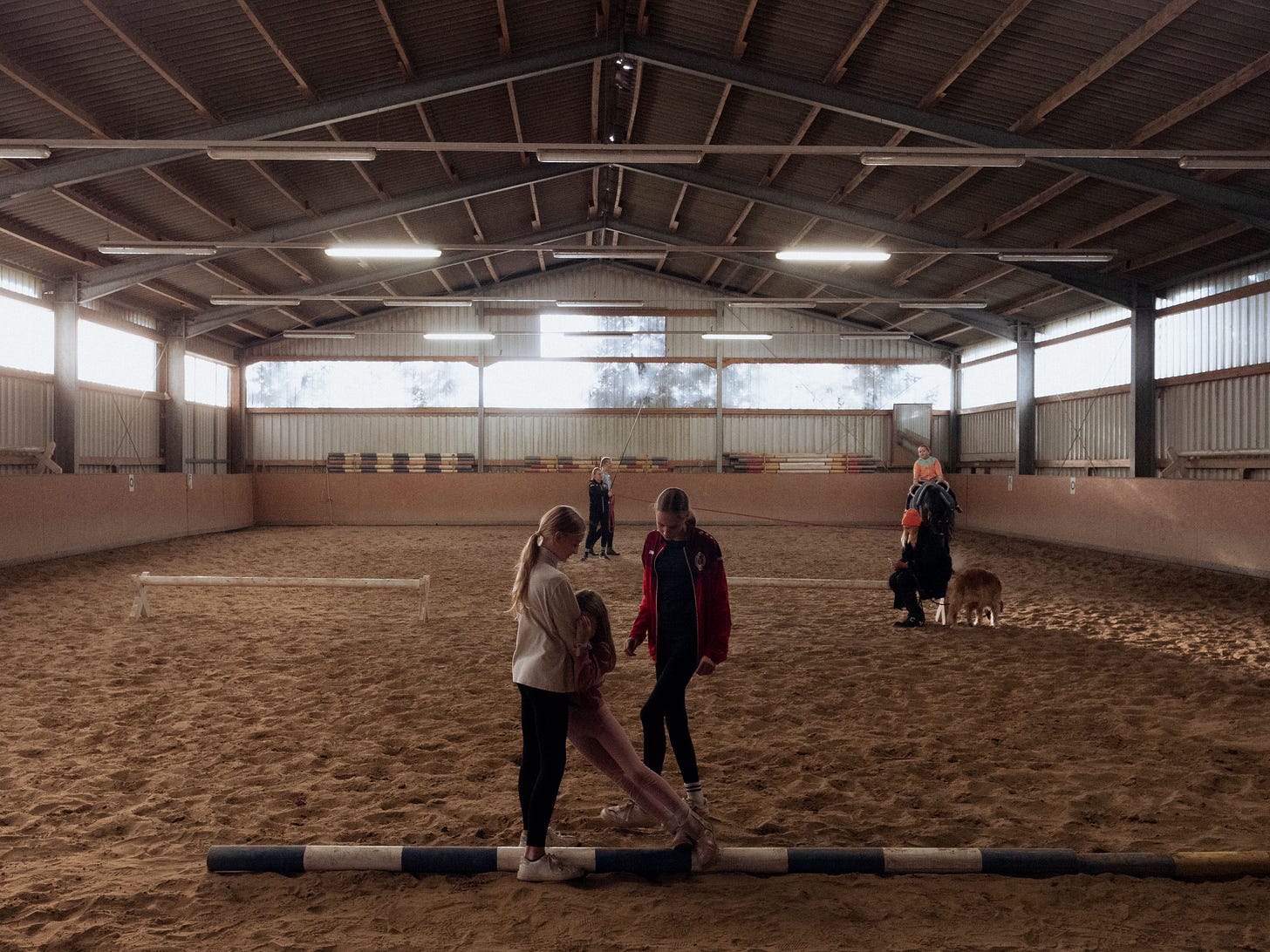 In einer Voltigierhalle ist das Training in vollem Gange. Man sieht im Vordergrund drei junge Voltigiererinnen, während im Hintergrund ein Pferd an der Longe läuft. In einer Voltigierhalle ist das Training in vollem Gange. Man sieht im Vordergrund drei junge Voltigiererinnen, während im Hintergrund ein Pferd an der Longe läuft.