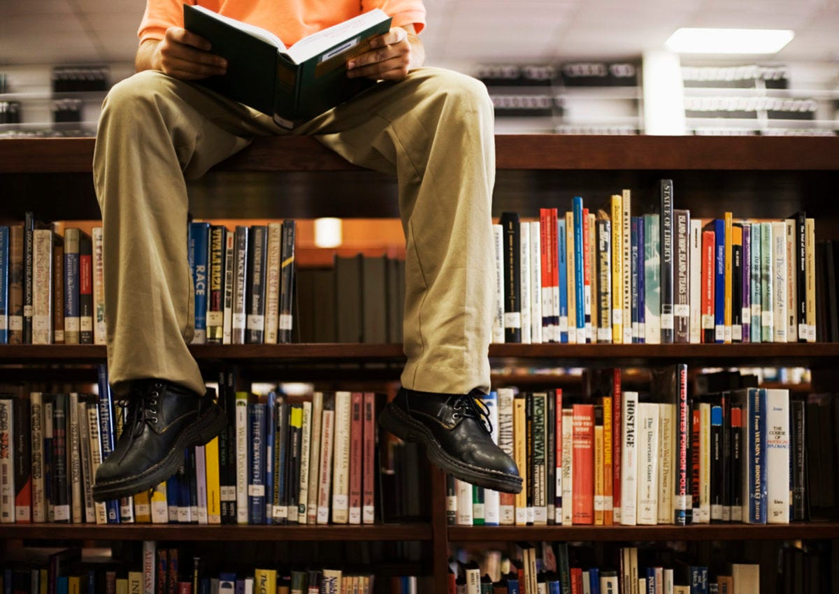 Man Reading Book and Sitting on Bookshelf in Library - Jeet Banerjee