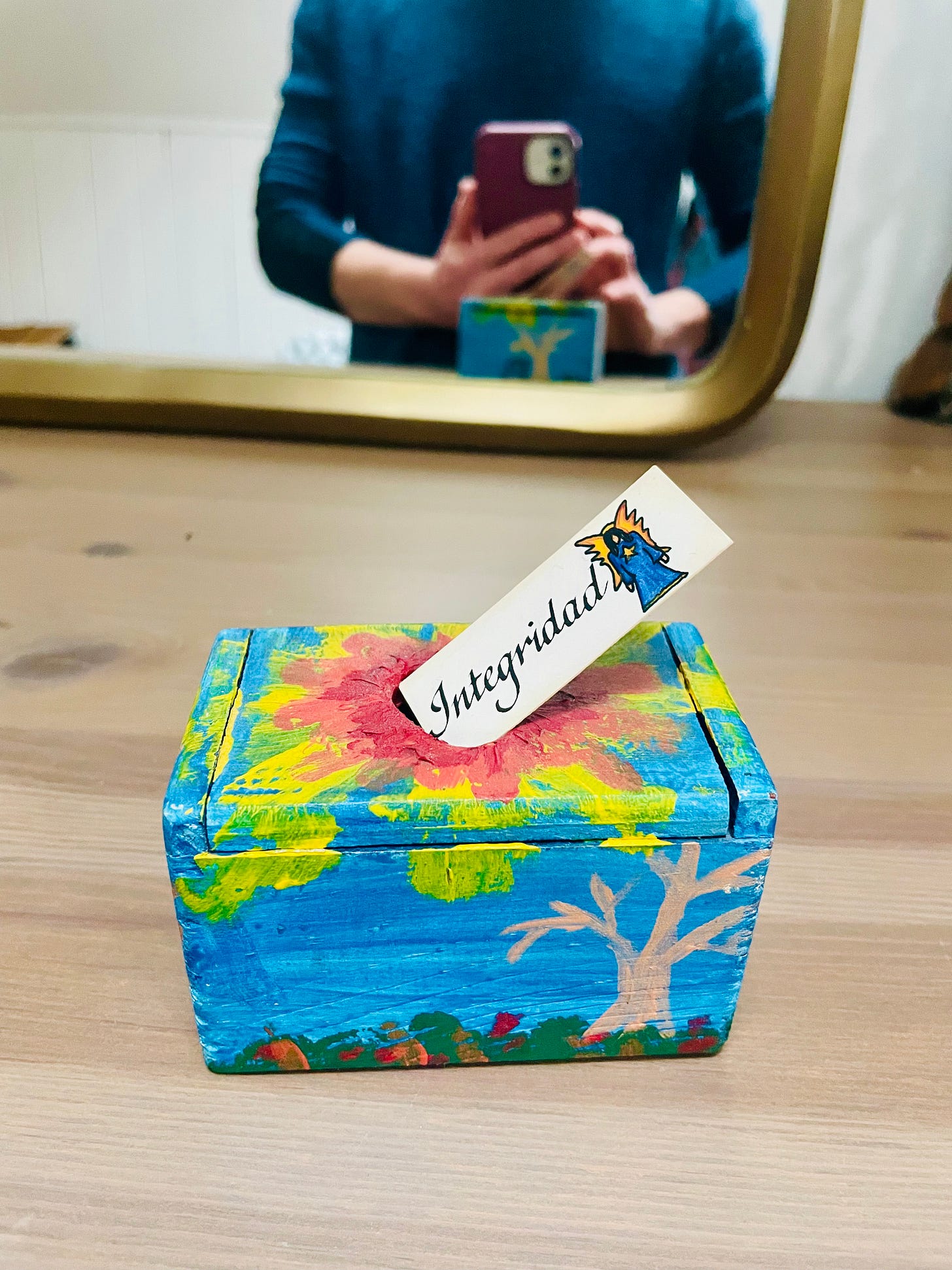 a blue pained box on a wood dresser, with a small white card (about 1 x 4 centimeters) with the word Integridad on it. In the background is a blurry mirror and a person taking a cell phone photo of the box.