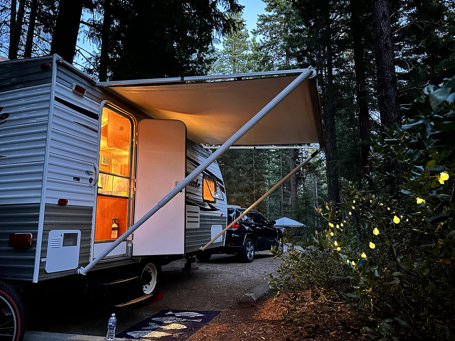 Small travel trailer at dusk with glowing light from inside the open door; canopy extended. A string of white lights on the bushes to the right of the photo adds to the coziness of the campsite.