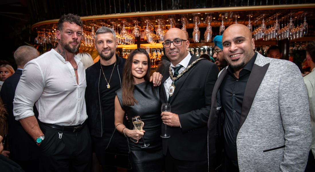A group of five people wearing white, black, and grey outfits holding glasses of champagne, stood in front of an orange-lit bar