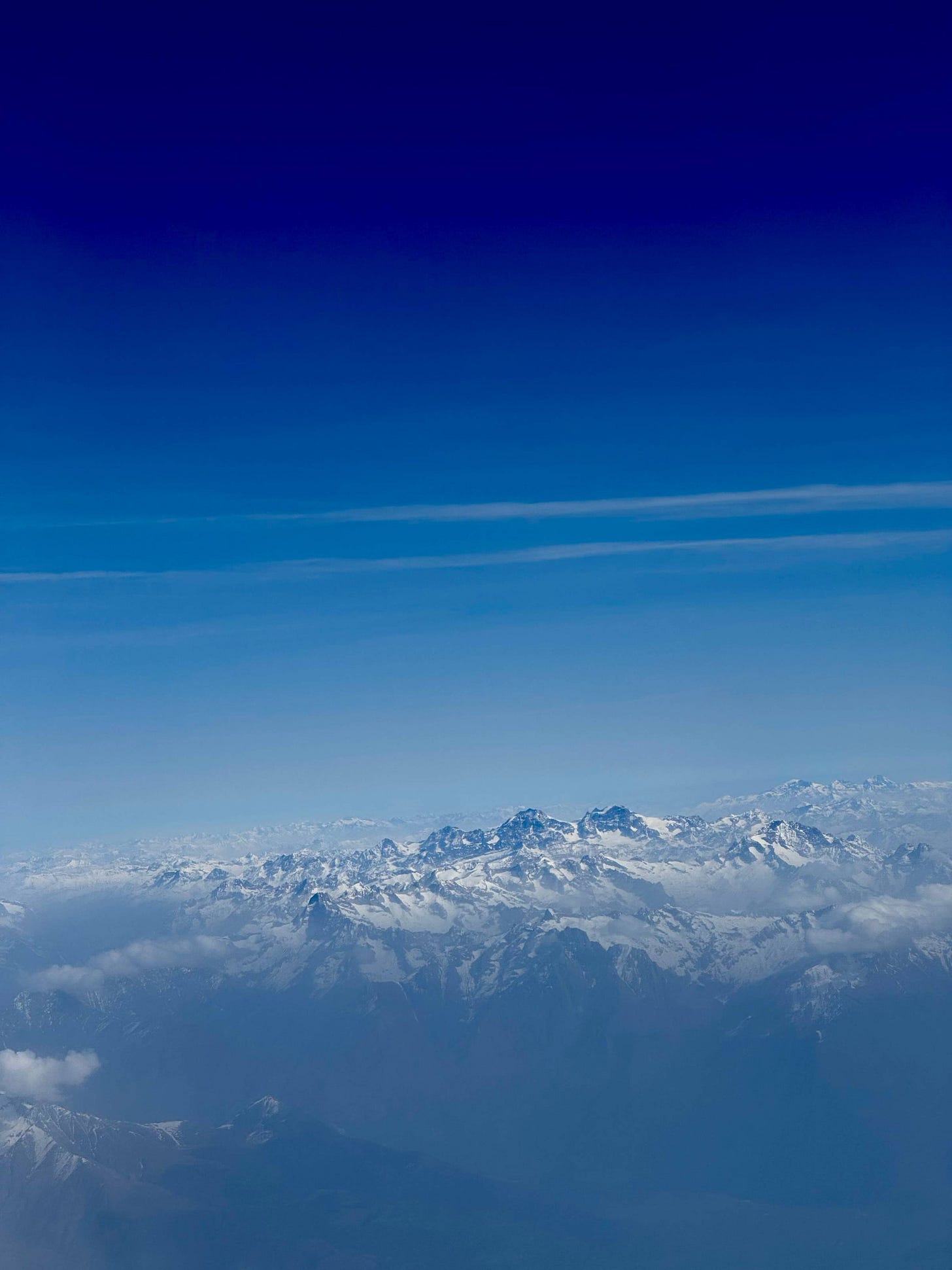 Aerial view of the snow-covered Alps from a plane window, peaks rising above cloud cover into a deep, gradient blue sky.