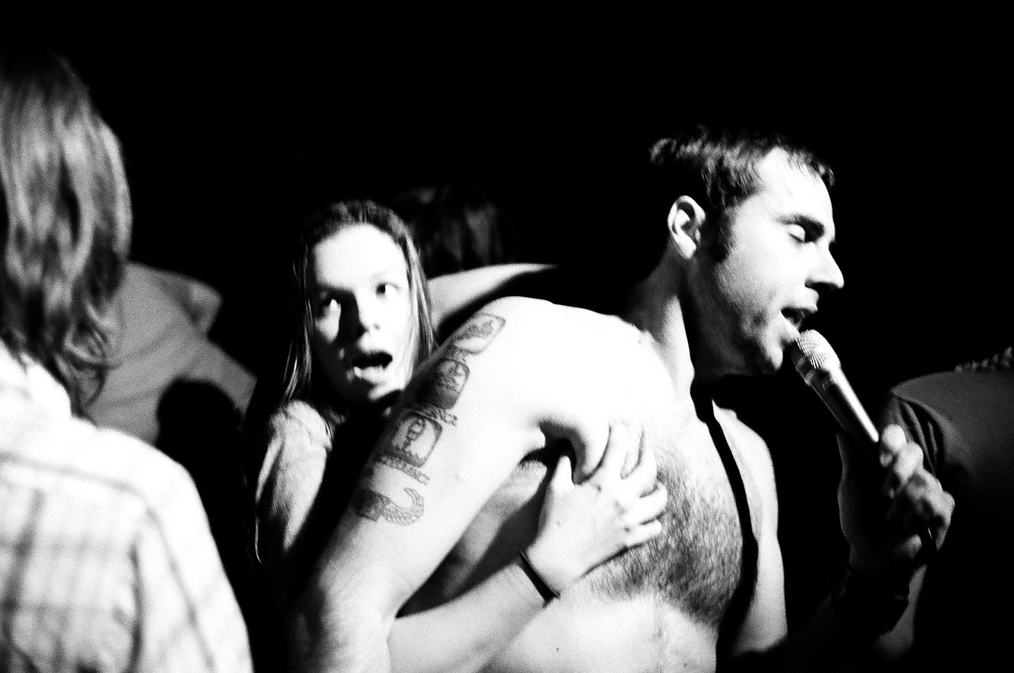 A slightly blurry photo of Derrick shirtless, singing into a microphone with his eyes closed. Amber stands behind him, jokingly grabbing his pecs while looking off to the side and making a shocked face. 