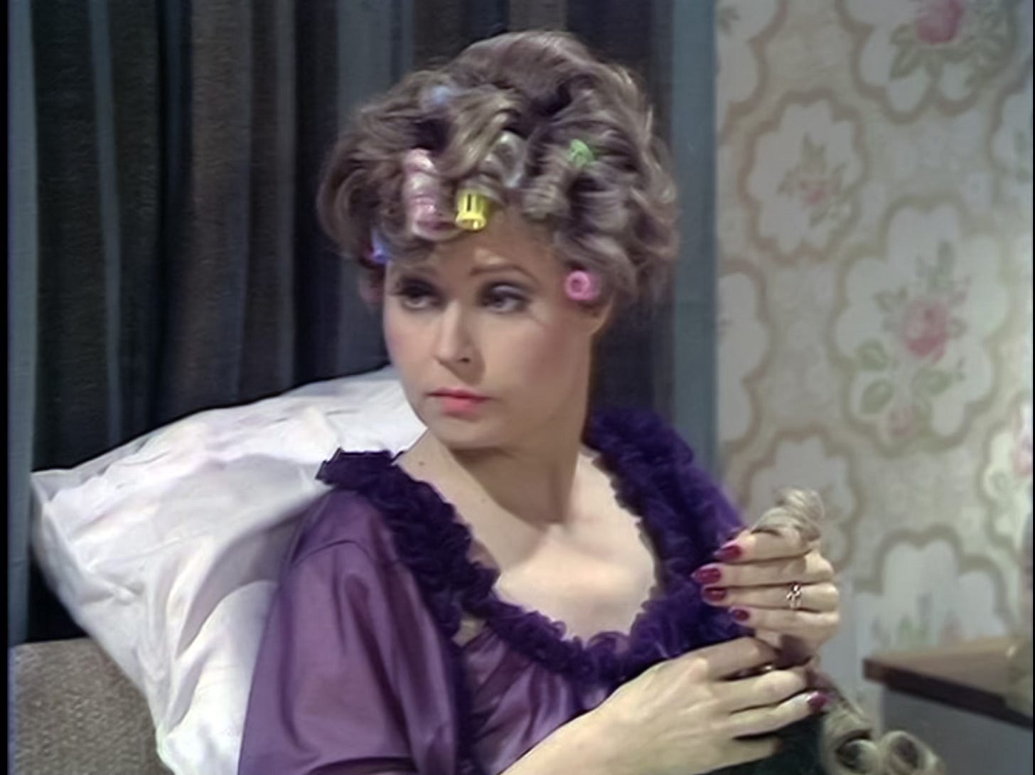 Sybil Fawlty sitting up in bed in curlers, fussing with a wig on her lap