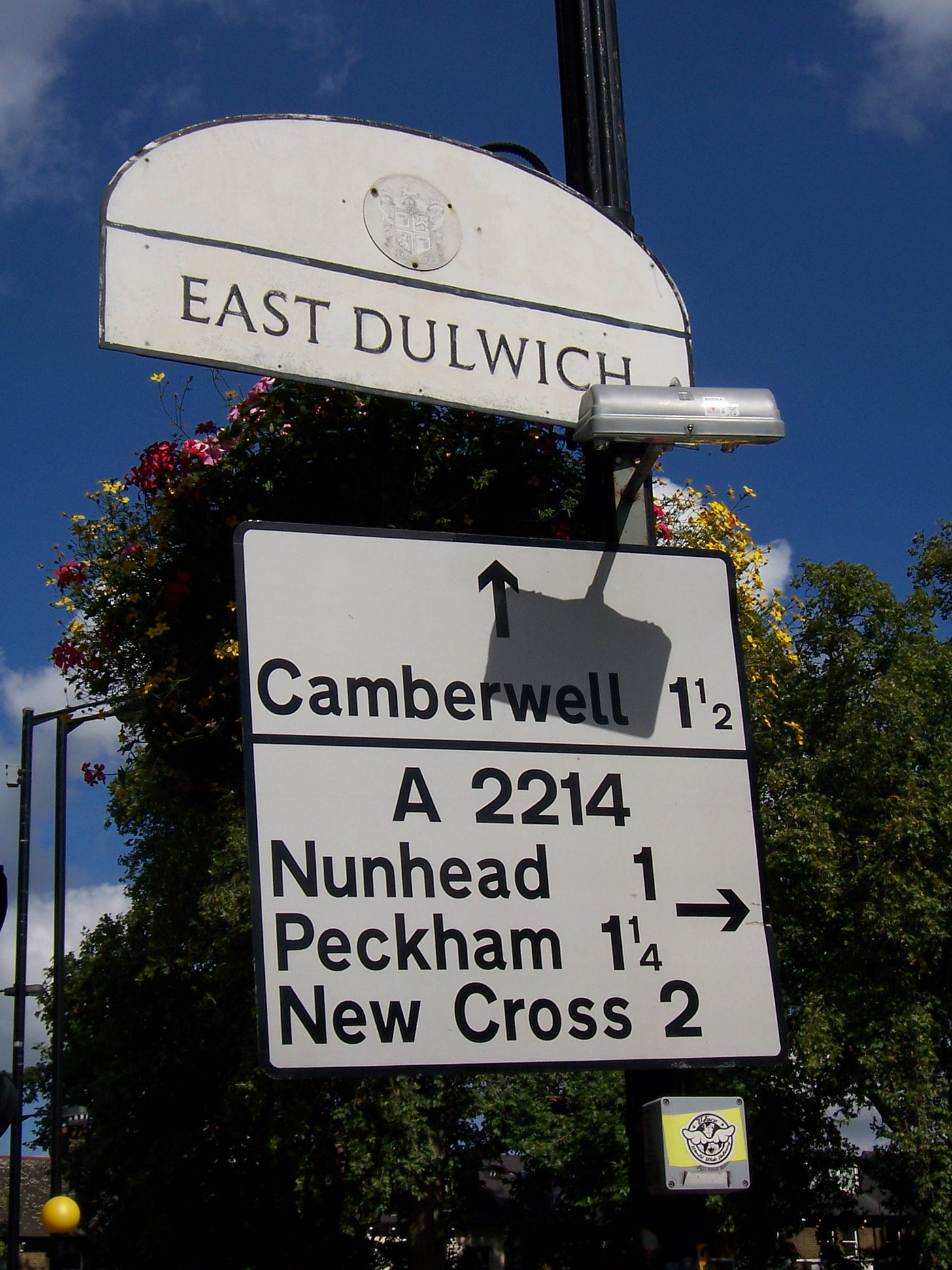Photograph of a road sign. A semi-circular panel at the top reads EAST DULWICH, while a much newer panel below it is mounted at a slight angle and reads Camberwell, A2214, Peckham, New Cross. Photograph of a road sign. A semi-circular panel at the top reads EAST DULWICH, while a much newer panel below it is mounted at a slight angle and reads Camberwell, A2214, Peckham, New Cross.