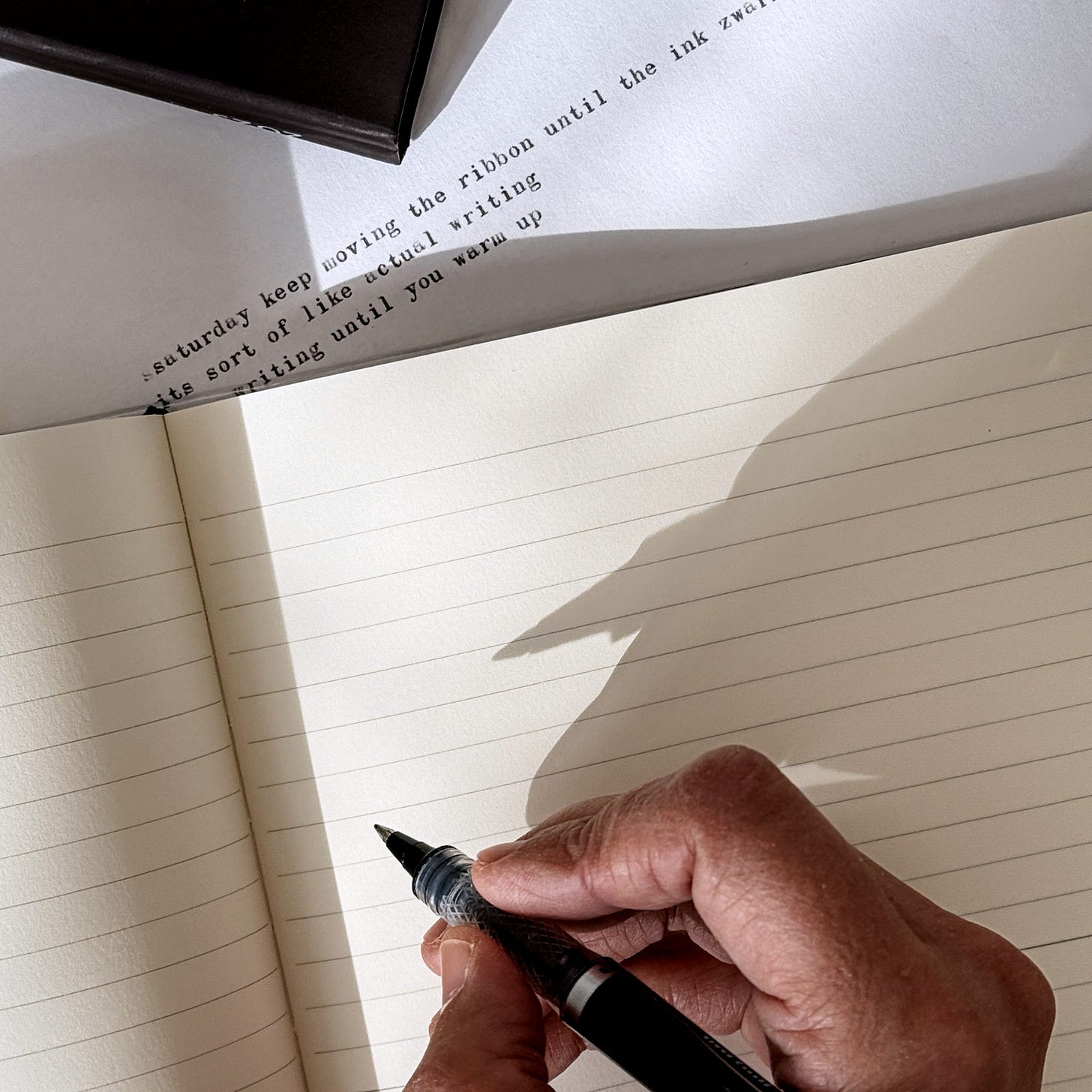 A hand poised to write in a lined notebook, lit by natural sunlight, with a Fabriano 2025 box and typewritten lines visible in the background.