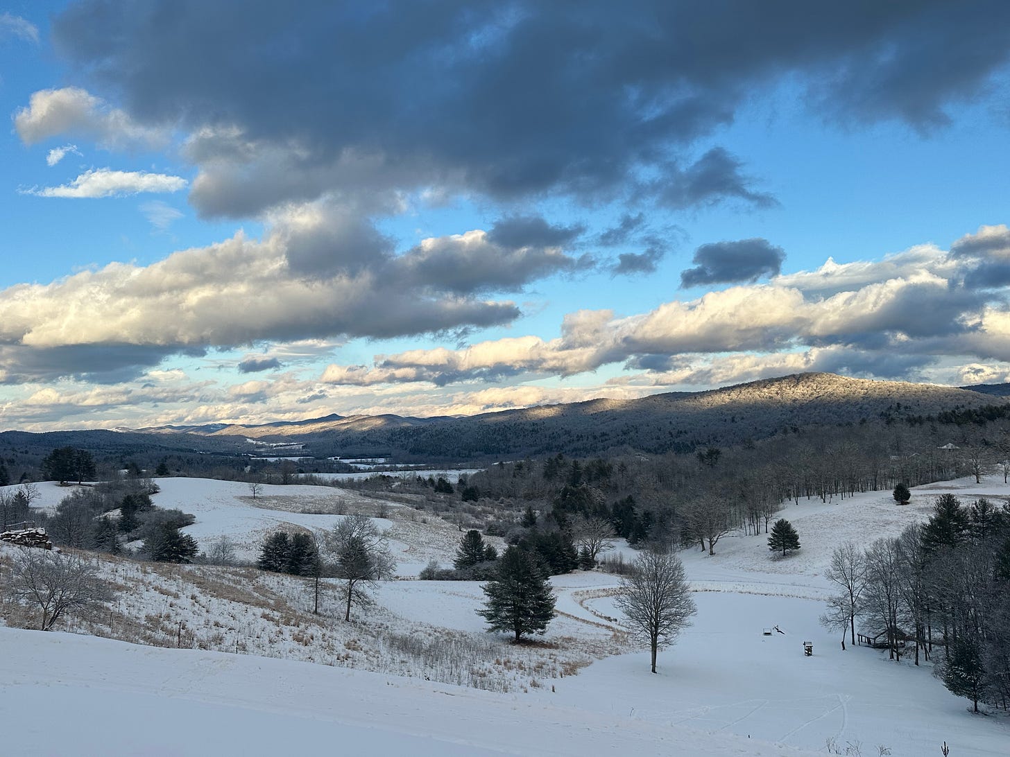 A photo of the appalachian mountains with snow in the foreground and a bright blue and grey clouded sky above. The sun shines through on parts of the mountains. 