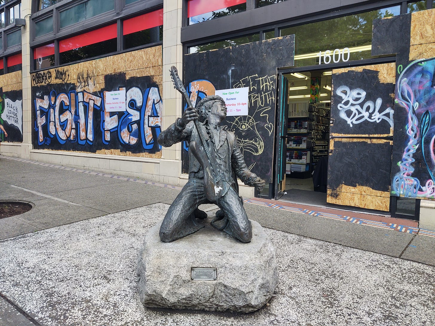 A bronze statue of Jimi Hendrix mounted on stone sits on the sidewalk in Capitol Hill. Jimi is on his knees playing his electric guitar. The Blick art supply store is behind the statue, the windows boarded up and graffiti'd with phrases including "Black Lives Matter." A bronze statue of Jimi Hendrix mounted on stone sits on the sidewalk in Capitol Hill. Jimi is on his knees playing his electric guitar. The Blick art supply store is behind the statue, the windows boarded up and graffiti'd with phrases including "Black Lives Matter."
