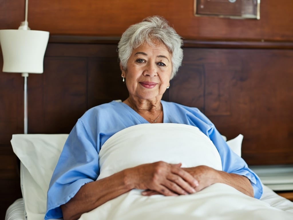 White woman with gray hair lying in hospital bed with her arms folded around the sheets that cover her up to her neck.