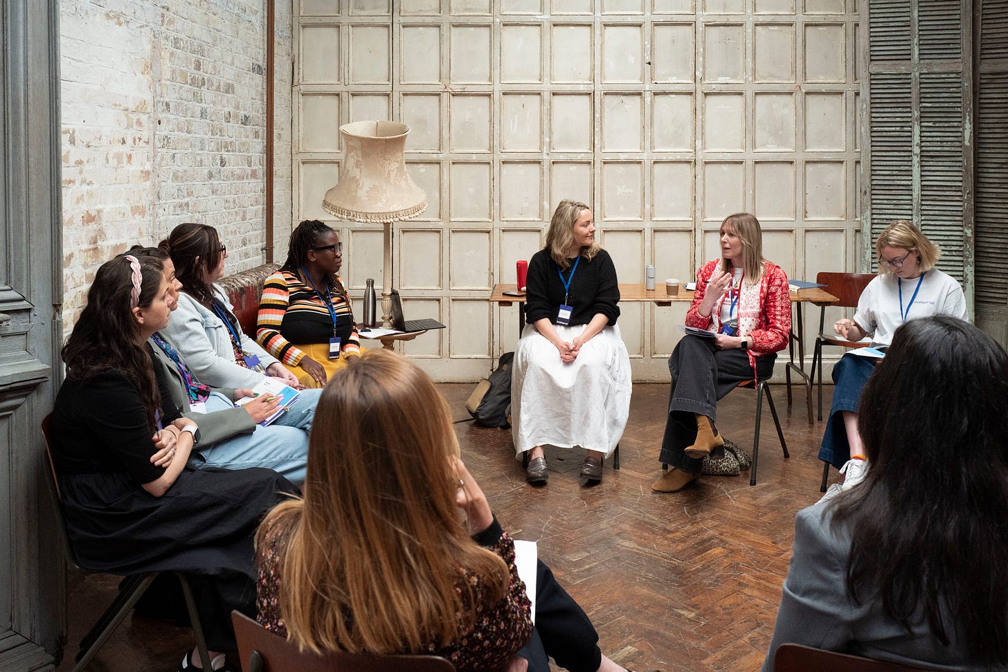 A vintage inspired room with whitewashed brick walls and a wooden floor hosting a workshop with Collette Philip and Ettie Bailey-King facilitating. Attendees look on as one person shares their feedback A vintage inspired room with whitewashed brick walls and a wooden floor hosting a workshop with Collette Philip and Ettie Bailey-King facilitating. Attendees look on as one person shares their feedback