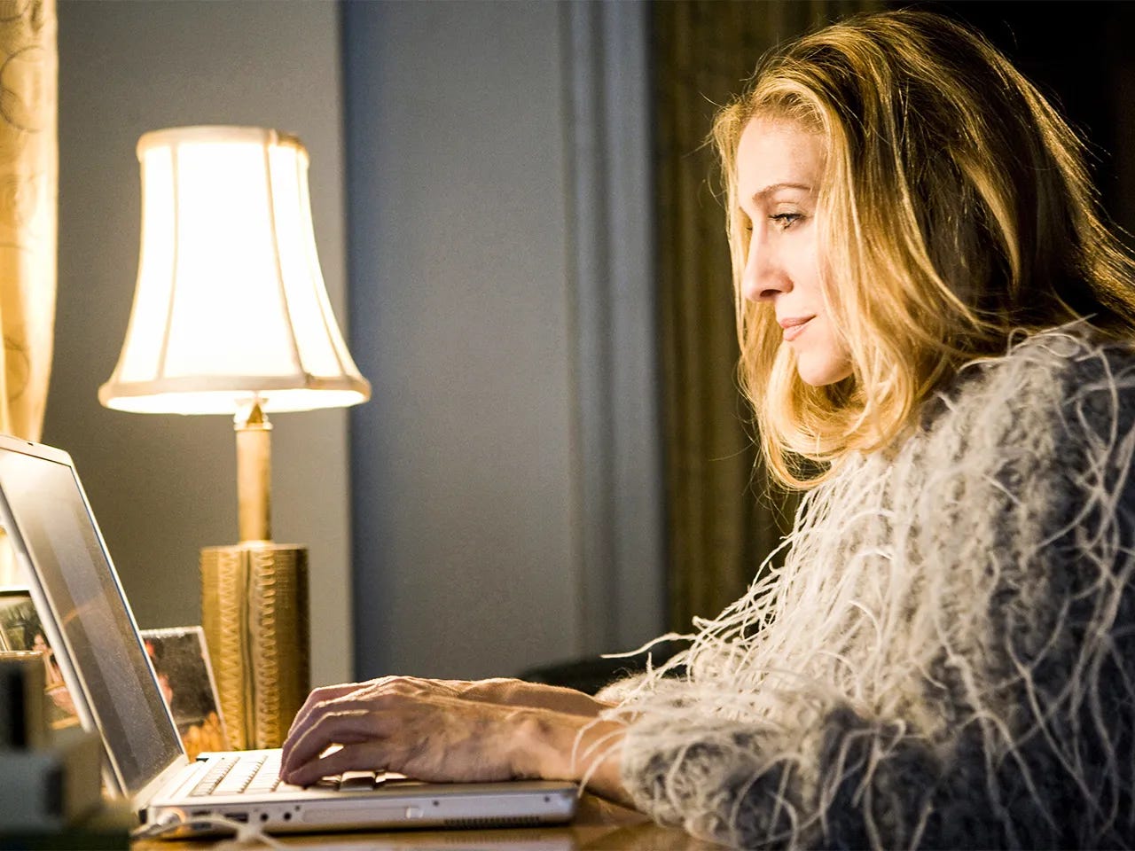 A side facing picture of Sarah Jessica Parker in the show Sex In the City as the character "Carrie" she writes at her laptop at a desk, wearing a feather coat, and with a lamp to the right of her laptop A side facing picture of Sarah Jessica Parker in the show Sex In the City as the character "Carrie" she writes at her laptop at a desk, wearing a feather coat, and with a lamp to the right of her laptop