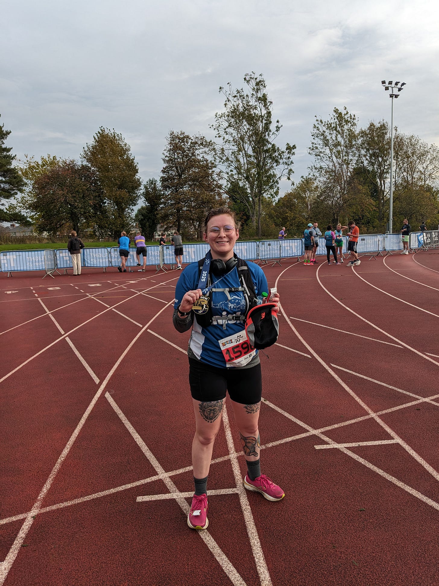 Ellen looking very red and sweaty standing with her running gear and holding a medal, smiling, with glasses on. She is on a running track. 