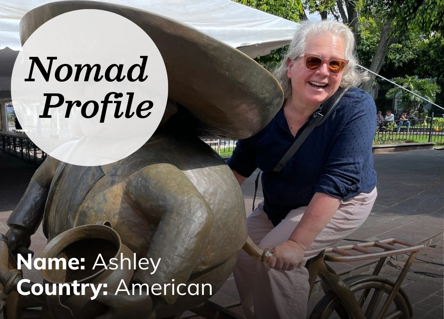 A white woman with shoulder-length grey hair is wearing a navy long-sleeved top and tan pants. She is wearing sunglasses, smiling, and riding a bicycle on a sunny day.