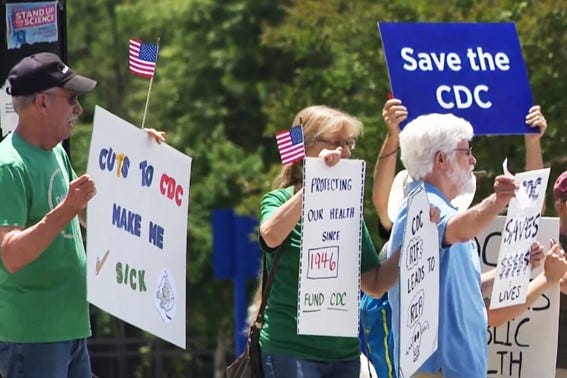 Group of protesters holding signs advocating for CDC funding, with messages like “Cuts to CDC Make Me Sick” and “Save the CDC,” while waving small American flags.