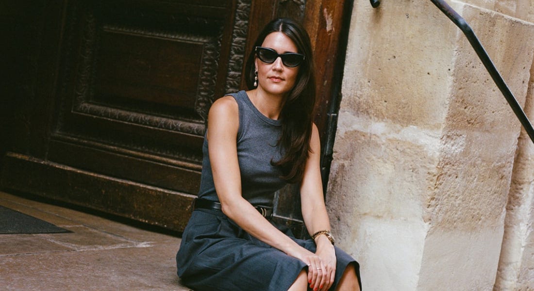 A woman - Bee Newham - wearing a grey top and shorts with sunglasses, sitting on a set of steps