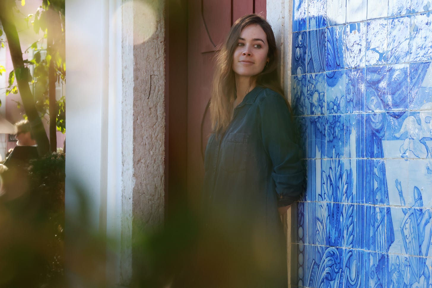 A photo of the author looking away from the camera to her left, leaning against a blue tile wall with medieval imagery painted on it in various shades of blue. She is smiling, but only slightly.